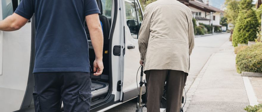 Ein Fahrer hält die Schiebetür auf, ein älterer Herr mit Rollator geht zum Wagen.