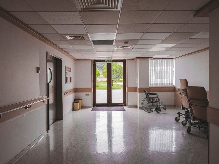 Heller Klinikflur mit Rollstuhl vor einer Glastür.