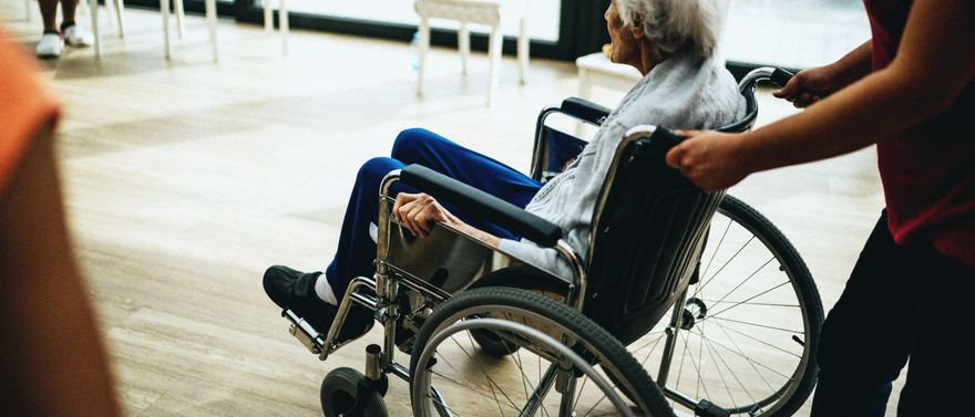 Eine Pflegekraft begleitet eine ältere Frau im Rollstuhl durch einen hellen Raum.