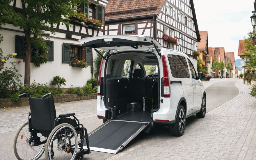 Weißer Kleinbus mit abgesenkter Rampe und bereitstehendem Rollstuhl in einer oberbayerischen Altstadtstraße.