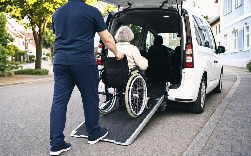 Ein Fahrer schiebt eine ältere Dame im Rollstuhl über die Heckrampe in einen weißen Van.