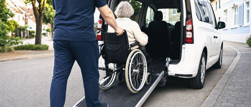 Ein Fahrer schiebt eine ältere Dame im Rollstuhl über die Heckrampe in einen weißen Van.